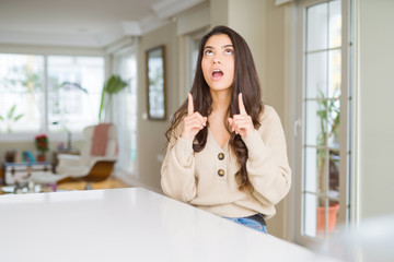Young beautiful woman at home amazed and surprised looking up and pointing with fingers and raised arms.