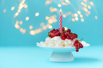 Pavlova birthday fruit cake with burn candle on pastel blue background against blurred lights. Selective focus. Healthy food or birthday celebration concept. Copy space
