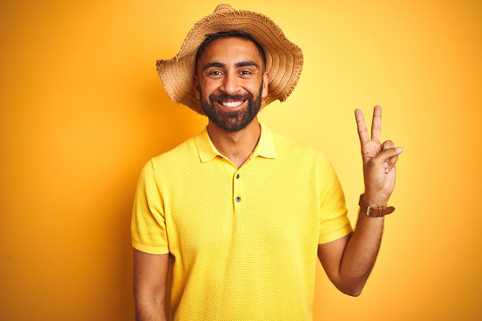 Young indian man on vacation wearing summer hat standing over isolated yellow background smiling with happy face winking at the camera doing victory sign. Number two.