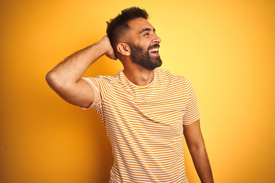 Young indian man wearing t-shirt standing over isolated yellow background smiling confident touching hair with hand up gesture, posing attractive and fashionable