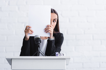 young lecturer suffering from fear of public speaking hiding face with paper sheet while standing on podium tribune