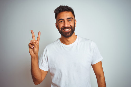 Young indian man wearing t-shirt standing over isolated white background smiling looking to the camera showing fingers doing victory sign. Number two.