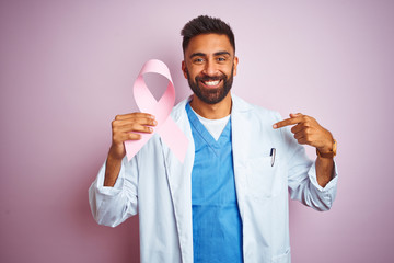 Young indian doctor man holding cancer ribbon standing over isolated pink background with surprise...
