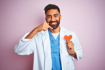 Young indian doctor man holding paper heart standing over isolated pink background very happy pointing with hand and finger