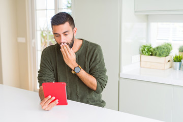 Handsome hispanic man using tablet at home cover mouth with hand shocked with shame for mistake,...