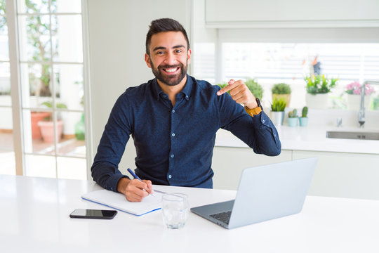 Handsome hispanic man working using computer and writing on a paper with surprise face pointing finger to himself