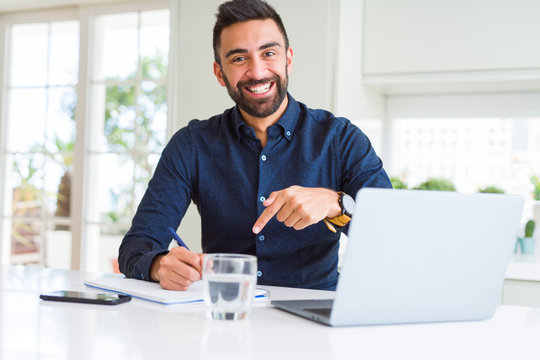 Handsome hispanic man working using computer and writing on a paper very happy pointing with hand and finger