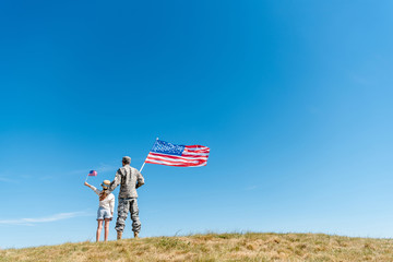 back view of kid in straw hat and military man holding american flags