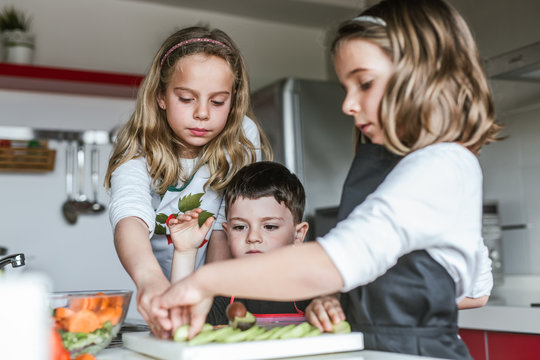 Little girls and boy cutting and peeling ripe vegetables while cooking healthy salad in kitchen together