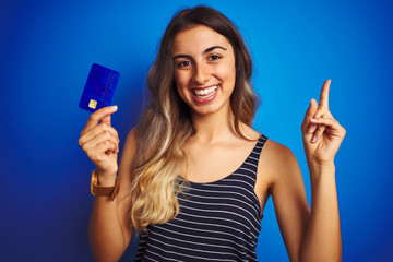 Young beautiful woman holding credit card over blue isolated background very happy pointing with hand and finger to the side