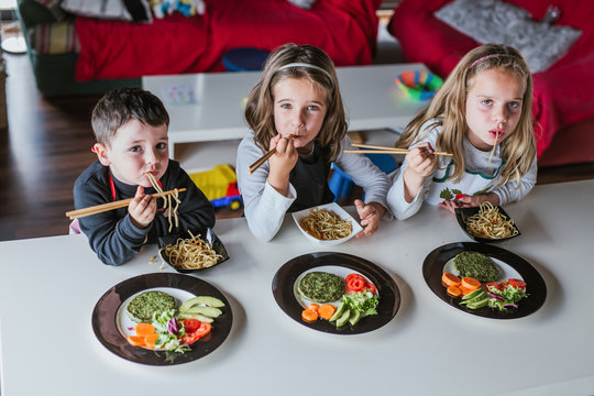 Boy and two girls looking at camera while enjoying tasty noodles with vegetarian cutlets and vegetables while sitting at table at home