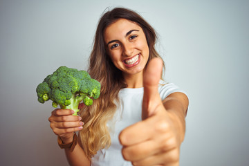 Young beautiful woman eating broccoli over grey isolated background happy with big smile doing ok sign, thumb up with fingers, excellent sign