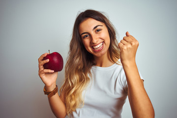 Young beautiful woman eating red apple over grey isolated background screaming proud and...