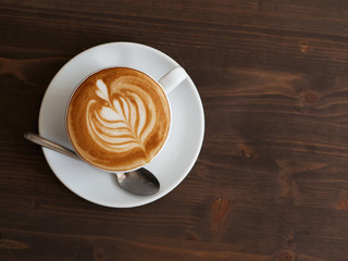 Overhead fresh brewed cappuccino coffee in white ceramic cup, saucer, spoon with beautiful handmade latte art on wooden table in cozy city cafe ready to drink