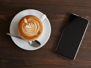 Overhead smartphone and fresh cappuccino coffee in white ceramic cup, saucer, spoon with beautiful handmade latte art on wooden table in cozy city cafe ready to drink
