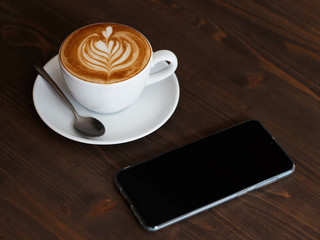 Modern smartphone and fresh brewed cappuccino coffee in white ceramic cup, saucer, spoon with beautiful handmade latte art on wooden table in cozy city cafe ready to drink, coffee break concept