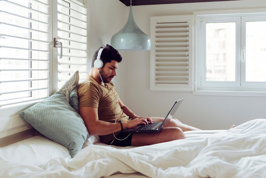 Young concentrated successful man in casual shirt and headphones sitting on bed using a laptop computer