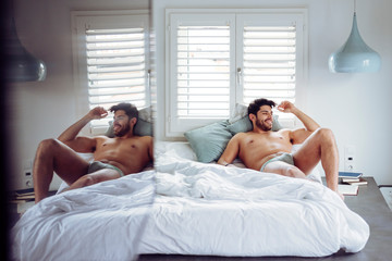 Young smiling successful sexy man with stylish hairstyle in grey briefs lying on bed at home and looking away
