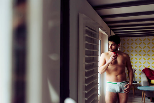 Side view of young pensive sexy handsome male in underwear standing near terrace at home and looking away