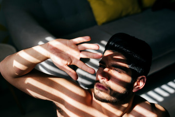From above young naked handsome male with stylish hairstyle standing near sofa and covering sunlight with hand