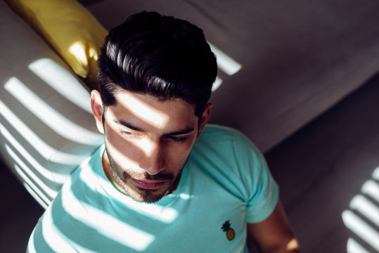 From above young tired depressed alone male in casual shirt sitting on sofa at home and looking at camera