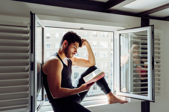 Side view of young serious handsome man in casual sleeveless shirt sitting on window and reading interesting book