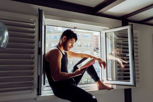 Side view of young serious handsome man in casual sleeveless shirt sitting on window and reading interesting book