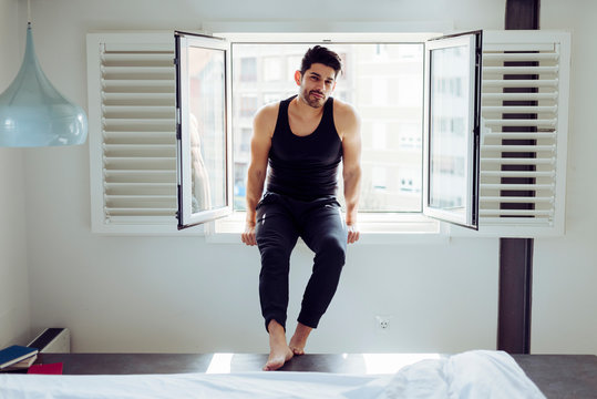 Young cheerful successful male in casual singlet sitting on window near bed with books and looking at camera