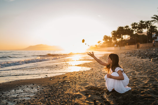 Side view of little girl in white dress sitting on beach and playing with stones on background of sundown