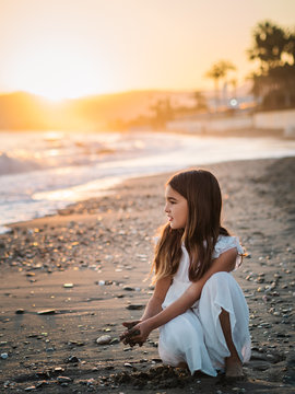 Cute pensive female kid in white dress playing with sand on seaside in sunlight