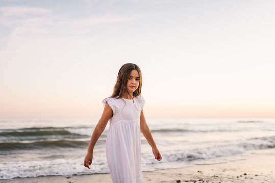 little girl in white dress walking and looking at camera on seashore on background of sunshine