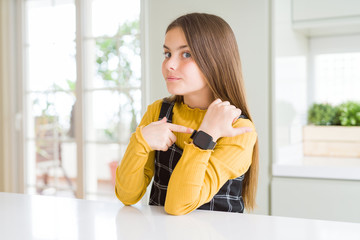 Young beautiful blonde kid girl wearing casual yellow sweater at home In hurry pointing to watch time, impatience, looking at the camera with relaxed expression