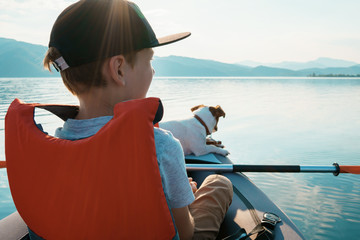 Happy boy with his dog Jack Russell Terrier paddling an inflatable kayak on the water mountain lake against the backdrop of beautiful orange sunset. Family sports vacation. Lens flare