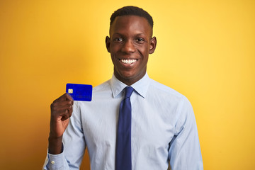 African american businessman holding credit card standing over isolated yellow background with a happy face standing and smiling with a confident smile showing teeth
