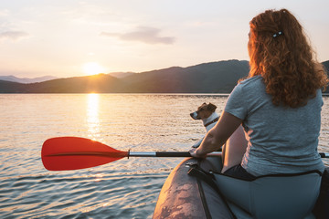 Happy young woman with her dog Jack Russell Terrier paddling on an inflatable kayak on the water of a large mountain lake against a beautiful orange sunset. Family Sports Weekend