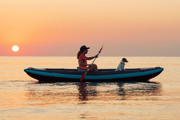 Red-haired young woman is rowing on an inflatable kayak by the sea with a dog Jack Russell Terrier on a background of pink sunrise in beautiful nature. Great disk of the rising sun. Sun rays. Sport