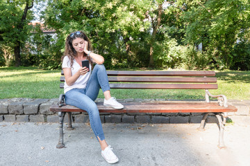 Online dating, Young beautiful girl sitting on the bench in the park looking on her phone waiting...