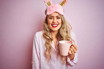 Young woman wearing pajama and mask drinking a cup of coffee over pink isolated background with a happy face standing and smiling with a confident smile showing teeth