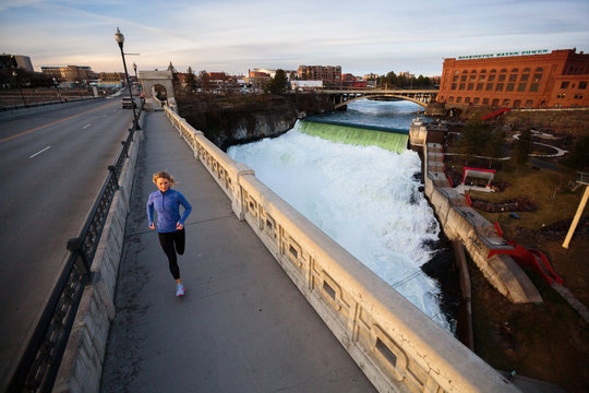 A blonde female runner runs on the Monroe Street Bridge adjacent a roaring Spokane Falls at high spring flow near downtown Spokane, Washington. The iconic Washington Water Power building is in the background.