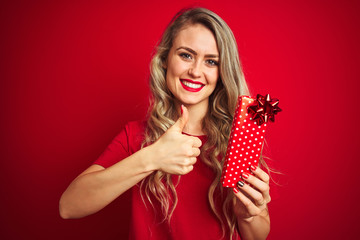 Young beautiful woman holding valentine gift over red isolated background happy with big smile doing ok sign, thumb up with fingers, excellent sign