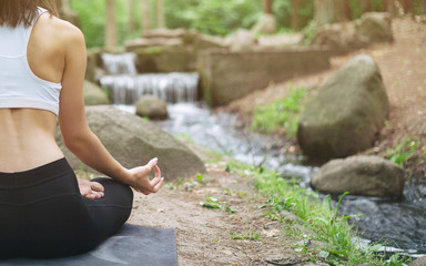 Young woman in yoga pose sitting near waterfall in sunny forest, Rear view. Copy space