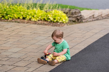 Horizontal side view of adorable fair toddler girl in summer clothes sitting in a driveway playing with a wooden pull toy