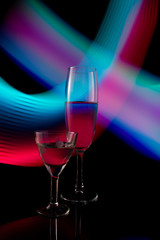 A two wine glass isolated on a black background with red, white, pink and orange neon light painting streaks of light behind them. Light wave, play of color, drawing with light.