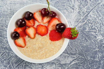 porridge in a white bowl with strawberries and cherries on a gray background
