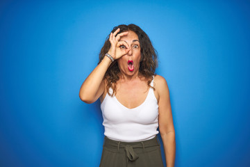 Middle age senior woman with curly hair standing over blue isolated background doing ok gesture shocked with surprised face, eye looking through fingers. Unbelieving expression.