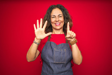 Middle age senior woman wearing apron uniform over red isolated background showing and pointing up with fingers number six while smiling confident and happy.