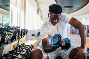 Young African American man sitting and lifting a dumbbell close to the rack at gym. Male weight training person doing a biceps curl in fitness center - obrazy, fototapety, plakaty