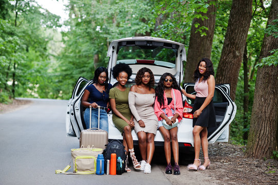 Group of five happy african american traveler girls sitting in car open trunk.