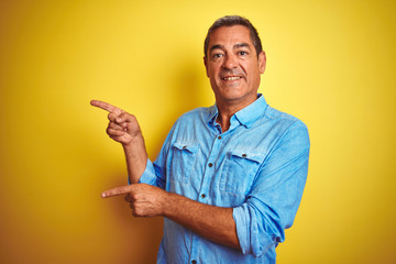 Handsome middle age man wearing denim shirt standing over isolated yellow background smiling and looking at the camera pointing with two hands and fingers to the side.