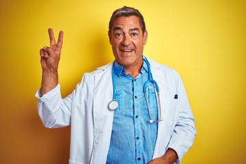 Handsome middle age doctor man wearing stethoscope over isolated yellow background smiling with happy face winking at the camera doing victory sign. Number two.